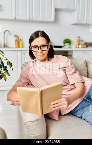 Eine reife Frau in kuscheliger Hauswäsche sitzt friedlich auf einer Couch, vertieft in ein Buch. Stockfoto