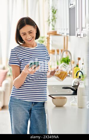 Reife Frau in gemütlicher Hauswäsche, die Handy in der Küche hält. Stockfoto