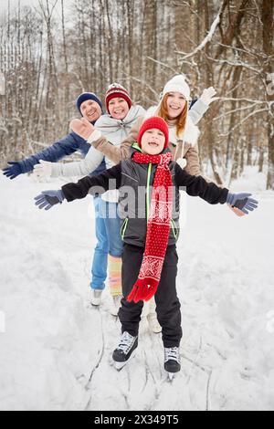 Glückliche vierköpfige Familie steht auf Schlittschuhen auf dem Eisweg im Winterpark und breitet Arme an die Seiten aus. Stockfoto