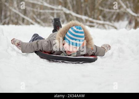 Kleiner Junge rutscht auf weichen Schlitten im Schnee im Winterpark. Stockfoto