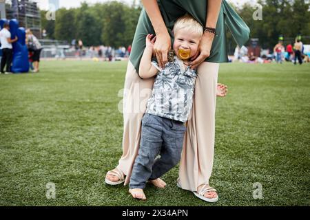 Kleinkind mit einer Puppe im Mund steht barfuß mit Mutterhänden auf grünem Gras. Stockfoto