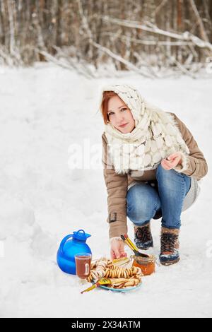 Teenager öffnet Glasgefäß mit Honig in der Nähe eines blauen Kannels, Glasbecher mit Getränk, Teller mit Pfannkuchen auf Schnee. Stockfoto