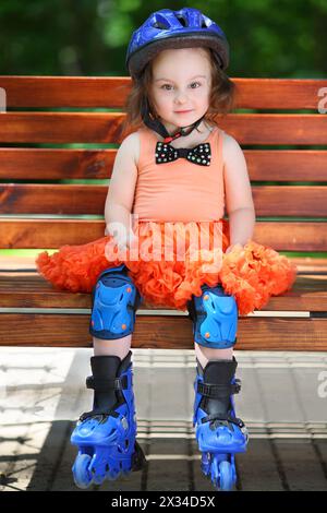 Das süße kleine Mädchen in Helm und Rollschuhen sitzt am Sommertag auf der Bank Stockfoto