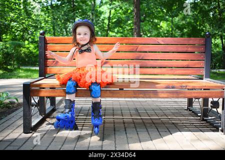 Niedliches kleines Mädchen mit blauem Helm und Rollschuhen sitzt am Sommertag auf der Bank Stockfoto