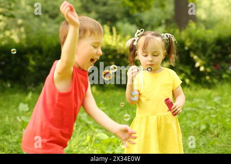 Das kleine Mädchen bläst Seifenblasen und ein glücklicher kleiner Junge fängt sie im Park Stockfoto