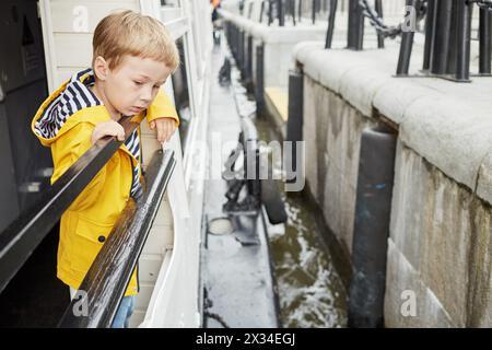 Kleiner Junge in gelber Jacke auf dem Deck des Vergnügungsbootes sieht das Boot, das sich dem Pier nähert. Stockfoto