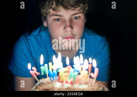 Der Junge sitzt in einem dunklen Zimmer und sieht brennende Kerzen auf der Geburtstagskuche an. Stockfoto