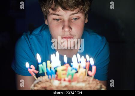 Der Junge sitzt in einem dunklen Zimmer und sieht brennende Kerzen auf der Geburtstagskuche an. Stockfoto