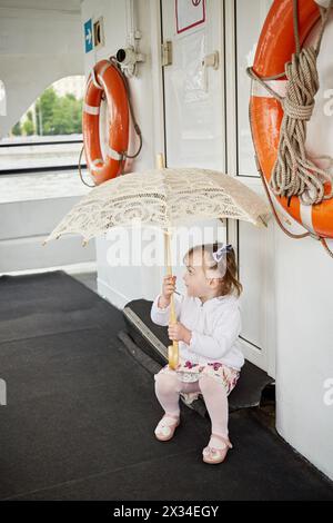 Ein kleines Mädchen mit Regenschirm sitzt auf der Tür auf dem Bootsdeck. Stockfoto