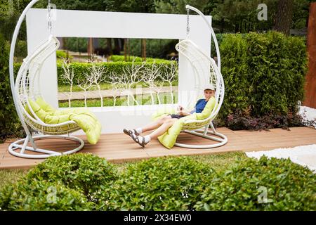 Ein lächelnder Teenager sitzt in einer weißen Schaukel und hängt an einer Kette im Sommerpark. Stockfoto