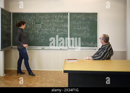 Frau spricht mit Zeiger nahe der Tafel mit Formeln und Mann hört ihr zu Stockfoto