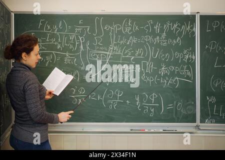 Der Lehrer spricht mit Buch und Zeiger in der Nähe der Tafel mit mathematischen Formeln im Schulunterricht Stockfoto
