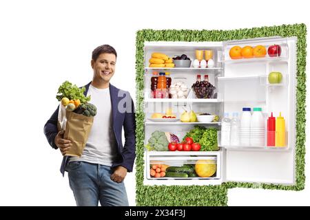 Junger Mann, der eine Lebensmitteltasche hält und sich auf einen grünen umweltfreundlichen Kühlschrank gestützt hat, der auf weißem Hintergrund isoliert ist Stockfoto
