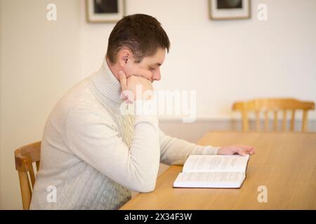 Ein Mann in weiß sitzt am Tisch, liest Buch und denkt im leeren Raum Stockfoto