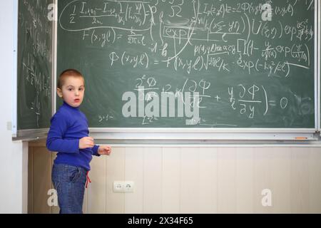 Der kleine süße Junge steht neben der Tafel mit Formeln im Klassenzimmer Stockfoto