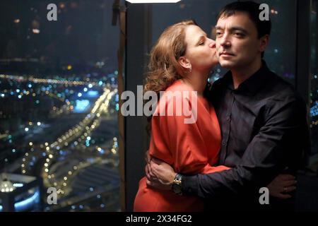 Eine Frau in rotem Kleid küsst abends in einem Restaurant einen Mann, der neben einem großen Fenster steht und einen Blick auf die Stadt hat Stockfoto