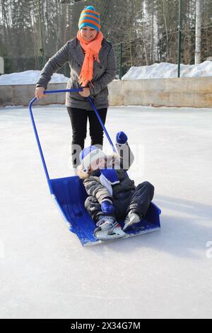 Mutter rollt den kleinen Sohn in einer großen Schneeschaufel auf einer Eislaufbahn Stockfoto