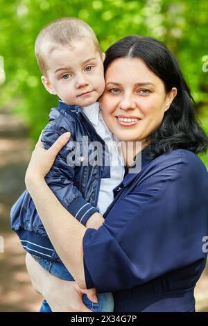 Die dunkelhaarige lächelnde Frau hält den kleinen Jungen in den Armen, der im Sommerpark steht. Stockfoto