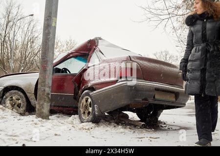Eine Frau im Wintermantel steht neben einem beschädigten Auto, das gegen die Laternenpfahl gestürzt ist. Stockfoto