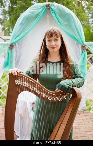 MOSKAU, RUSSLAND - 30. MAI 2015: Junge Harfenspielerin der Musikband Polca an Ri hält Arpa gegen Arch dekoriert grün und wh Stockfoto