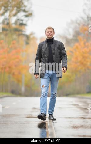 Junger Mann in schwarzer Jacke und blauer Jeans, der durch die Gasse im Park läuft Stockfoto