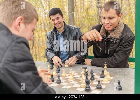 Drei junge Männer in schwarzen Jacken sitzen im Pavillon, zwei von ihnen spielen Schach, konzentrieren sich auf den richtigen Mann Stockfoto