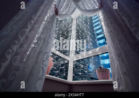 Fenster mit Vorhängen und leuchtender Girlande in dunklem Raum, geringer Blickwinkel. Stockfoto