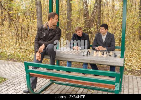 Drei junge Männer sitzen im Pavillon zum Schachspiel im Herbstpark Stockfoto