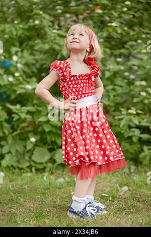 Das kleine Mädchen in rotem, gepunktetem Kleid steht mit den Händen akimbo und blickt nach oben auf den grasbewachsenen Rasen. Stockfoto