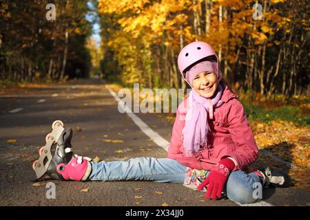 Glückliches Mädchen in einem Schutzhelm sitzt ausgestrecktes Bein in den Rollen auf dem Asphalt im Herbstpark Stockfoto
