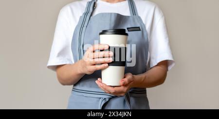 Eine Barista mit Schürze hält eine große wiederverwendbare Kaffeetasse vor sich. Stockfoto