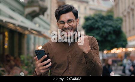 Glücklicher hoffnungsvoller indischer arabischer ethnischer Mann Mann Geschäftsmann männlicher junger Student hält Handy-Smartphone und wünscht Hoffnung betend gekreuzt Stockfoto