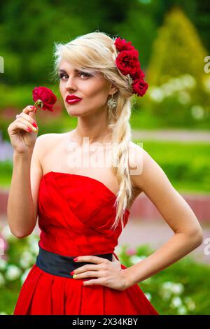 Halblanges Porträt einer schönen hübschen Frau in rotem Kleid mit Rose in den Händen im Sommerpark Stockfoto