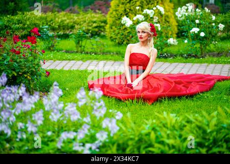 Halblanges Porträt einer schönen Frau in rotem Kleid im Sommerpark, die auf Gras zwischen Blumen sitzt Stockfoto