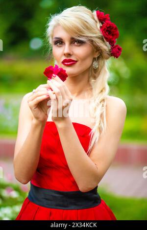 Halblanges Porträt einer hübschen Frau in rotem Kleid, die im Sommerpark nach Blumen riecht Stockfoto