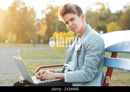 Junger gutaussehender Mann, der bei Sonnenuntergang auf Einer Bank im Park sitzt. Hält Ein Notebook in der Hand und arbeitet. Stockfoto