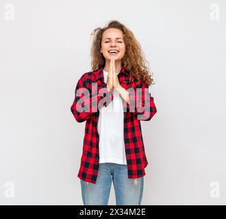 Lockige Frau in einem roten Hemd drückt Emotionen der Freude und Erwartung vor einem hellgrauen Hintergrund aus und gestikuliert sie. Stockfoto