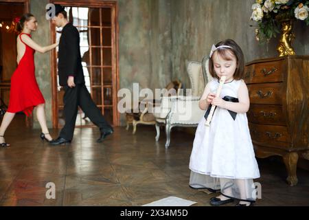 Kleines Mädchen im Kleid spielt Flöte im Zimmer mit tanzendem Paar, konzentriert sich auf das Kind Stockfoto