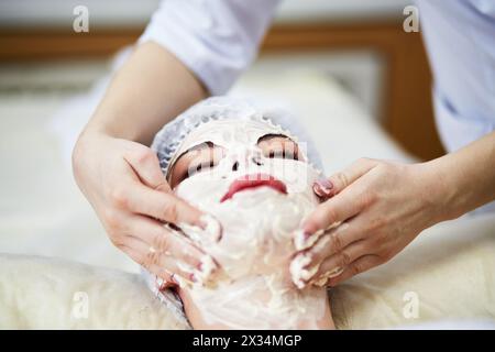 Beautician smears cosmetic mask with hands on face and neck of woman lies on couch in beauty salon. Stockfoto