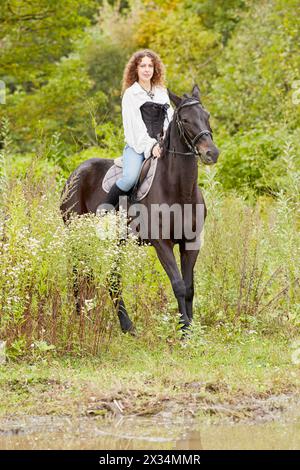 Lächelnde Frau mit lockigen Haaren reitet auf einem Bay-Pferd im Park. Stockfoto