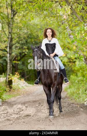 Lächelnde Frau mit lockigen Haaren reitet auf einem Bay-Pferd im Park. Stockfoto