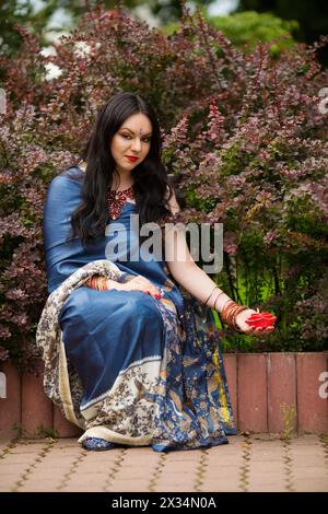 Brünette in blauem Sari sitzt neben Berberitze mit roter brennender Kerze in Form von Rosen Stockfoto