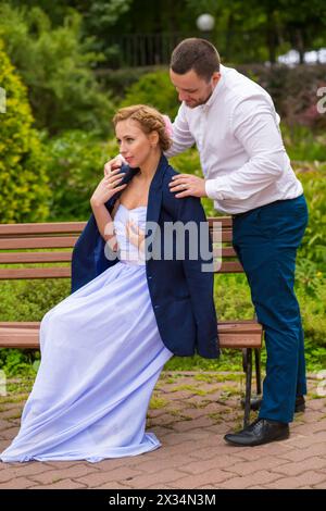 Hübsche Frau sitzt auf der Bank, der Mann zieht seine Jacke für Mädchen in den grünen Sommerpark Stockfoto