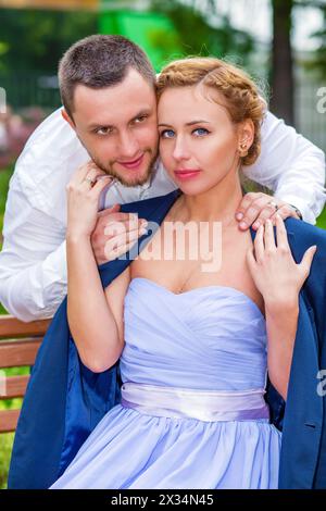 Frau in Kleid und Jacke sitzt auf der Bank und Mann umarmt sie im Sommerpark Stockfoto