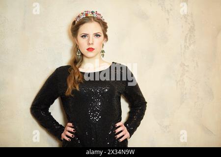 Eine junge Frau im dunklen Strickkleid steht mit ihren Armen akimbo an der Wand im Studio. Stockfoto