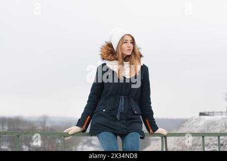 Niedliches Mädchen In Wintermütze Und Jacke, Das Auf Dem Geländer Sitzt. Im Winter Im Freien. Stockfoto