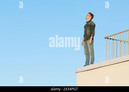 Ein junger Mann in Denim-Kleidung steht am Rand des Gebäudedachs vor blauem Himmel. Stockfoto