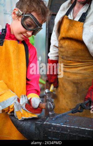 Mädchen in Brille und gelber Schürze mit Hammer, der Amboss in Schmiede schlägt Stockfoto