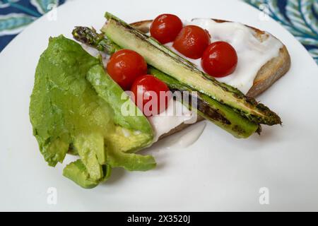 Gegrillter Spargel mit Kirschtomaten und Avocado auf Sauerteigtoast mit einer Schicht Blumenkohlmousse. Alles auf einem weißen Teller auf einem Blatt serviert Stockfoto