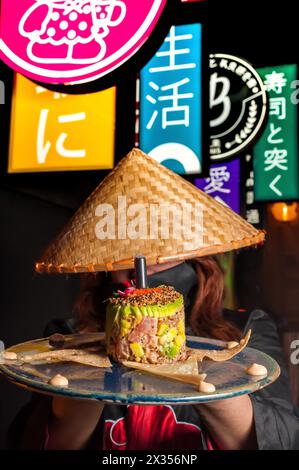 Rothaarige in japanischer Verkleidung und konischer Hut, der einen Lachsartar mit hellen Werbelichtern hinter ihr serviert Stockfoto